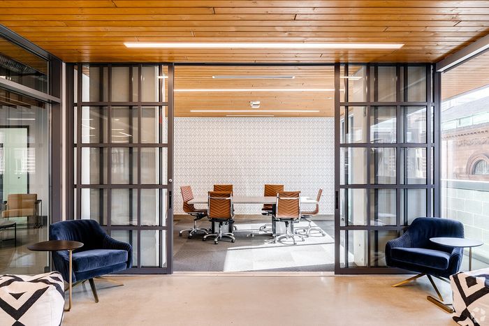 Contemporary office space featuring a glass-enclosed conference room with modern brown leather chairs, stylish blue velvet armchairs, and elegant lighting.