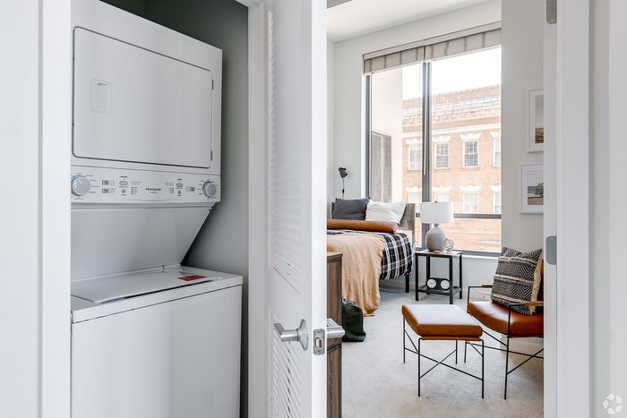 Modern apartment featuring an in-unit stacked washer and dryer, with a cozy bedroom and large window for natural light.