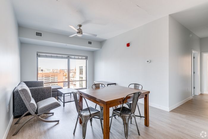 Bright and modern apartment living space with large windows offering city views, sleek wooden dining table, and stylish gray lounge chairs.