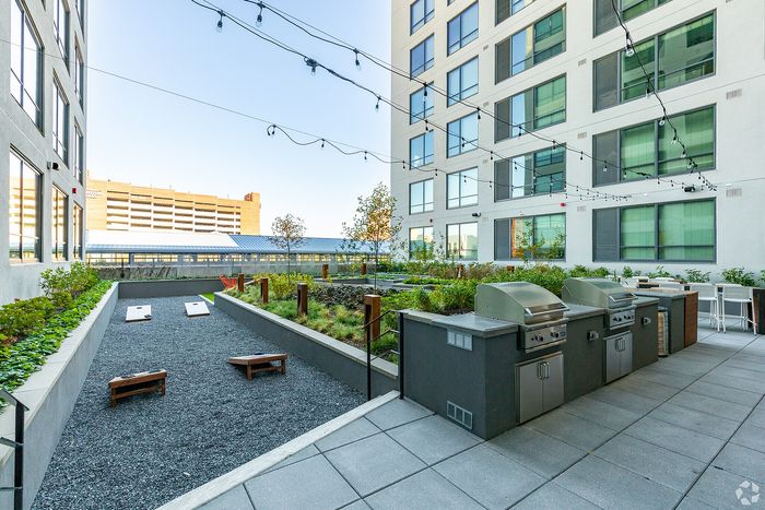 Modern apartment rooftop with lush greenery, outdoor grills, and cozy seating areas under string lights.