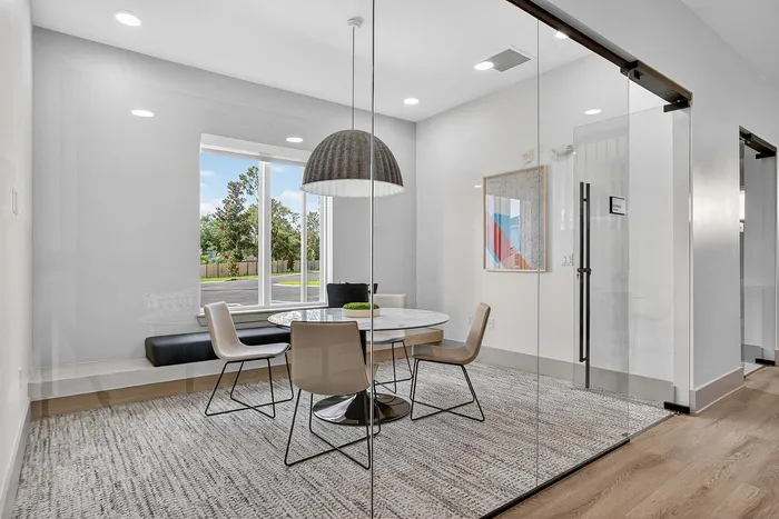 Modern glass-walled dining area with chic round table, stylish chairs, and abundant natural light from large window, offering a serene view.