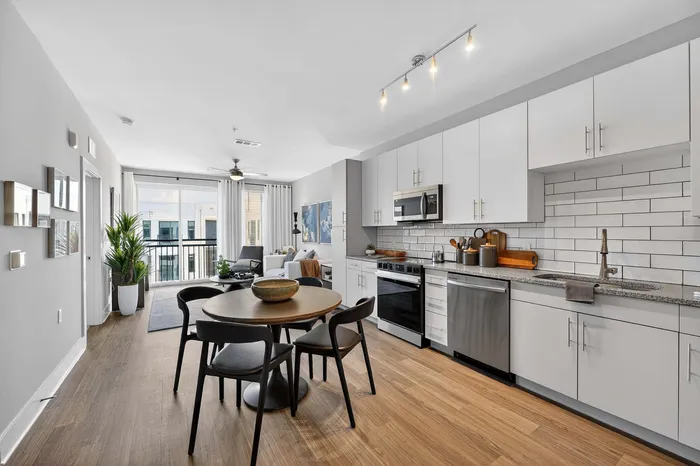 Bright, modern open-plan kitchen and living space with sleek white cabinetry, wooden floors, and natural light pouring in through large windows leading to a balcony.