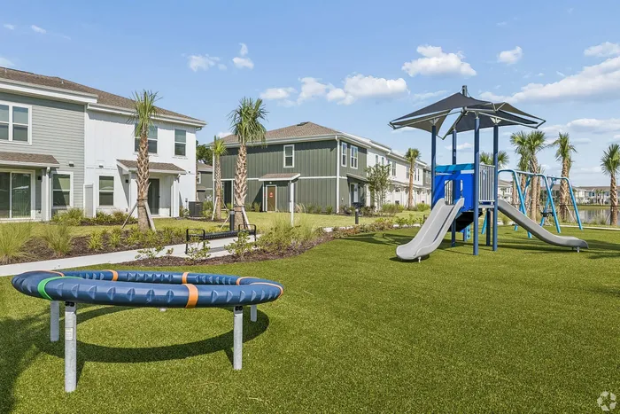 Modern residential community featuring lush green lawns, palm trees, and a children's playground with slides and swings under blue skies.
