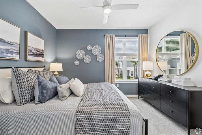 Chic bedroom with elegant decor, featuring a plush bed adorned with patterned cushions, stylish wall art, and a generous window inviting natural light. A sleek dresser with modern accents completes the tranquil ambiance.