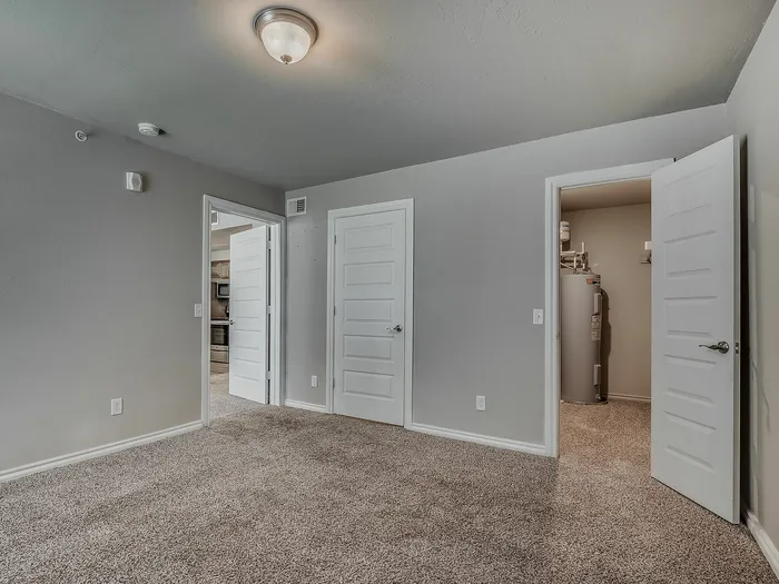 Spacious and modern bedroom with plush carpeting, leading to an open kitchen view, featuring neutral walls and stylish lighting.