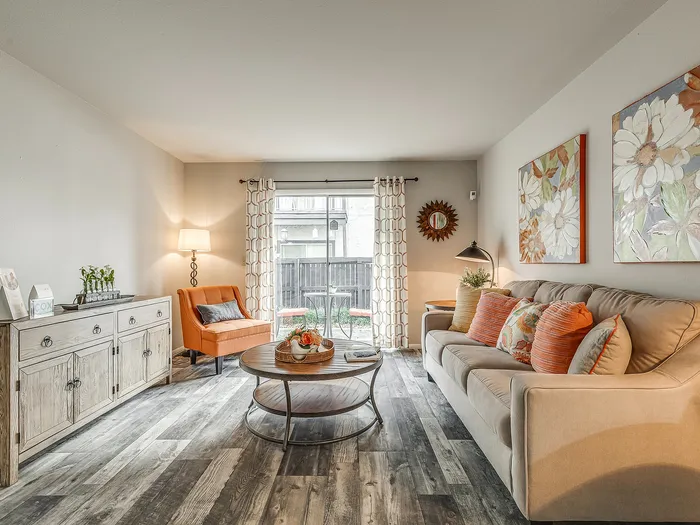 Inviting living room with elegant decor, featuring a cozy beige sofa, vibrant orange armchair, artistic floral paintings, and abundant natural light from large windows.