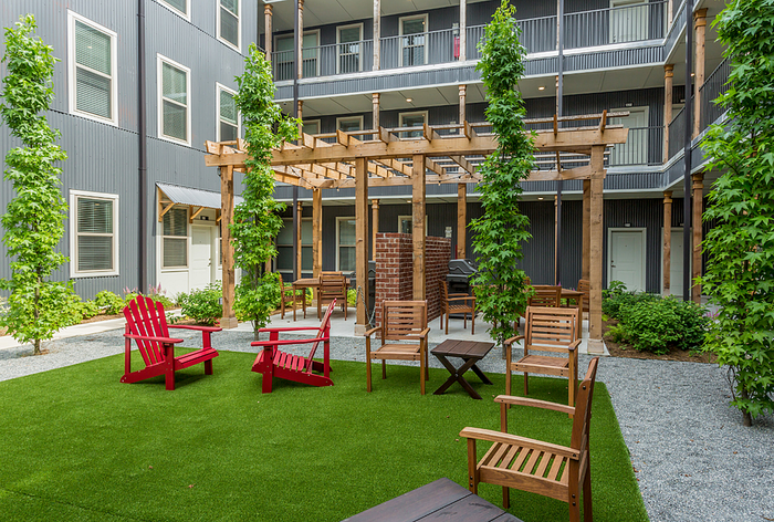 Charming courtyard oasis featuring lush greenery, stylish wooden pergola, and inviting seating area with vibrant red and brown chairs.