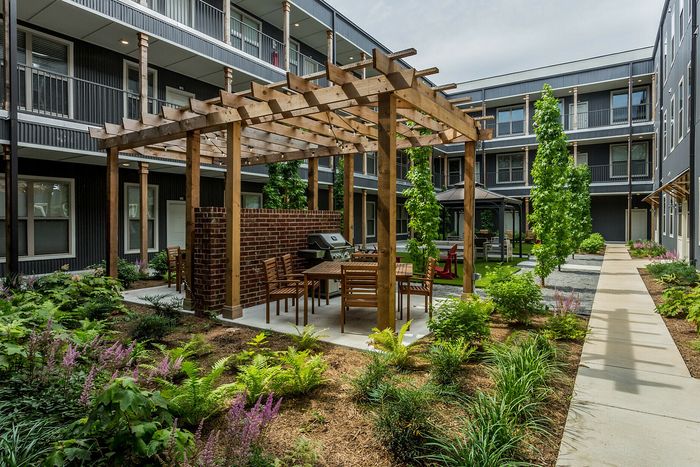 Modern apartment courtyard featuring a wooden pergola with seating, lush green landscaping, and elegant brick accents.