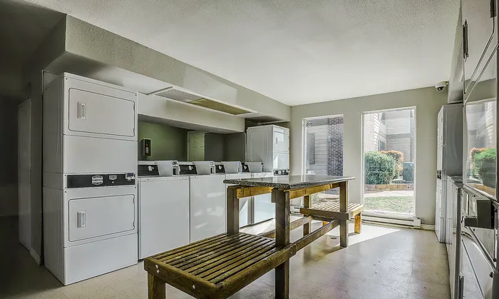 Bright apartment laundry room featuring modern washing machines, cozy seating with natural light, and view of lush garden.