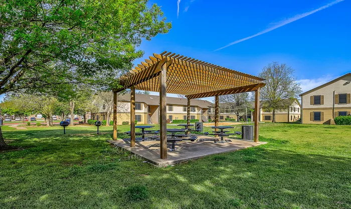 Charming outdoor space featuring a wooden pergola with picnic tables, surrounded by lush green grass and nearby residential buildings.