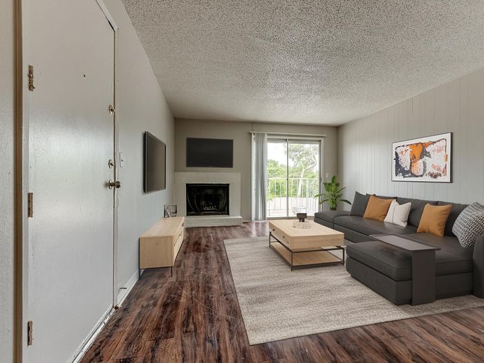 Inviting living room with warm wooden flooring, stylish gray sectional sofa, and modern decor. Features a cozy fireplace and large windows offering abundant natural light and green views.
