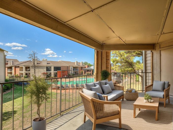 Inviting apartment balcony with stylish wicker furniture, offering a stunning view of a community pool and charming residential buildings under a clear blue sky.