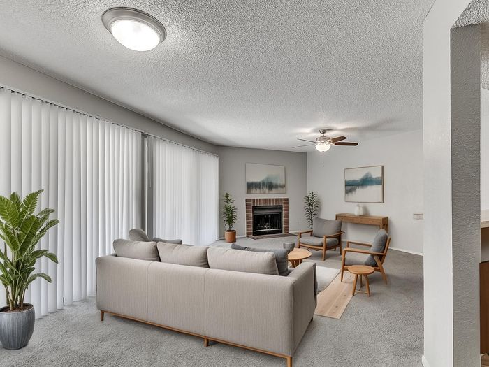 Bright and inviting living room with modern furniture, large windows with vertical blinds, and a cozy fireplace, accented by tasteful artwork and lush green plants.