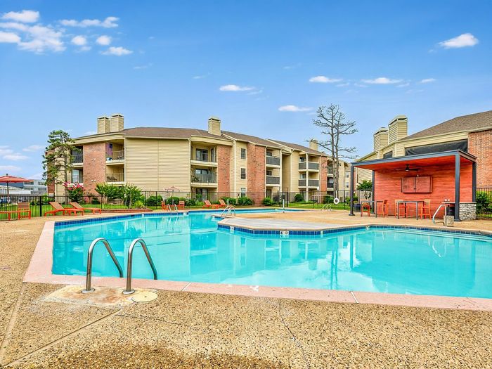 Vibrant apartment complex with a sparkling outdoor pool, surrounded by lush green landscaping and comfortable lounge chairs for relaxation.
