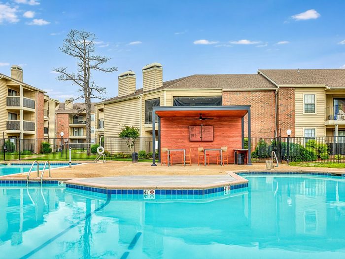 Inviting apartment complex featuring a sparkling swimming pool, surrounded by elegant buildings and lush greenery under a clear blue sky.