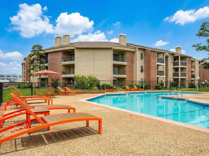 Charming apartment complex with a sparkling pool, surrounded by vibrant orange loungers and lush greenery under a bright blue sky.