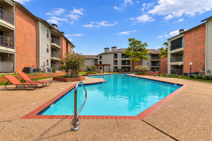 Inviting apartment complex with a sparkling outdoor pool surrounded by colorful sun loungers and beautiful greenery under a clear blue sky.