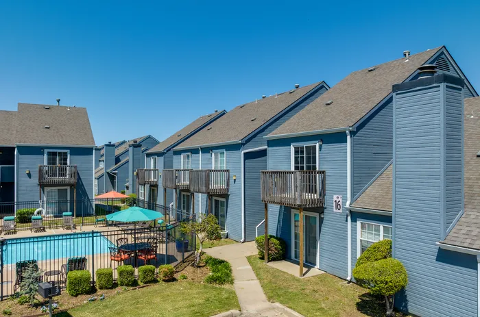Charming blue townhomes with private balconies, overlooking a pristine pool area complete with sunlit seating and vibrant umbrellas.