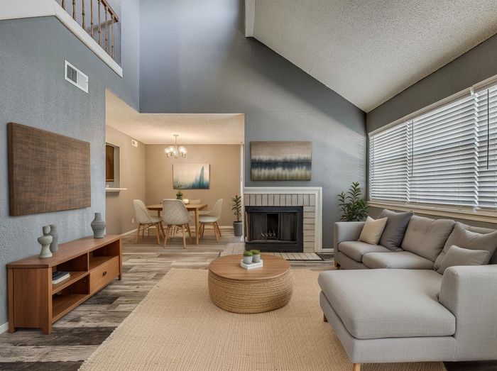 Elegant living room with a cozy gray sofa, large windows, and a modern fireplace, leading to a stylish dining area with chic artwork and contemporary furniture.
