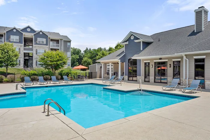 Inviting apartment complex featuring a sparkling outdoor swimming pool, modern lounge chairs, and charming architecture set against lush greenery.