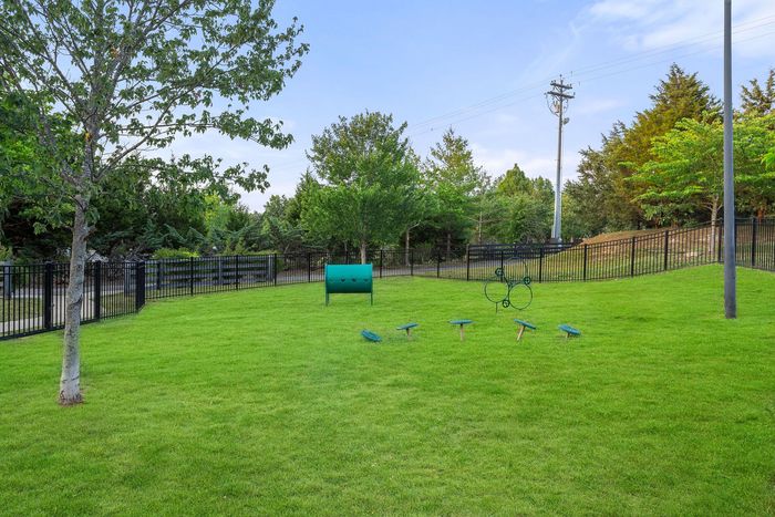 Vibrant grassy playground enclosed by a secure iron fence, featuring interactive green play equipment surrounded by lush trees.