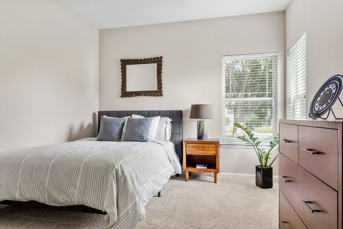 Bright and inviting bedroom with a stylish gray upholstered bed, natural light streaming through large windows, modern decor, and a cozy atmosphere.