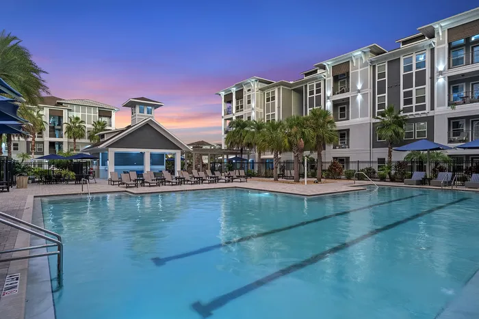 Luxurious apartment complex with a large pool, surrounded by palm trees and stylish lounge chairs, under a vibrant sunset sky.