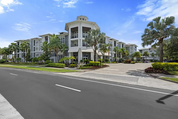 Luxurious modern apartment complex surrounded by lush palm trees and vibrant landscaping, set against a clear blue sky.