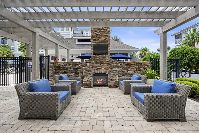 Inviting outdoor patio featuring cozy wicker furniture with blue cushions, a charming stone fireplace, and lush greenery under a stylish pergola.