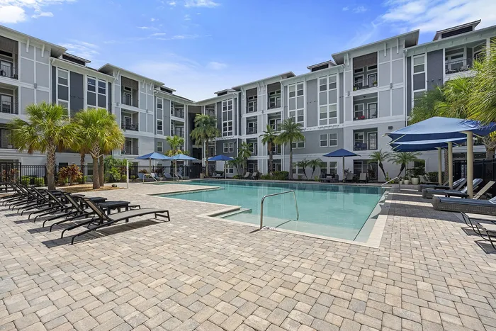 Luxurious apartment complex with modern design featuring a spacious pool area, surrounded by lush palm trees, elegant lounge chairs, and shaded umbrellas.