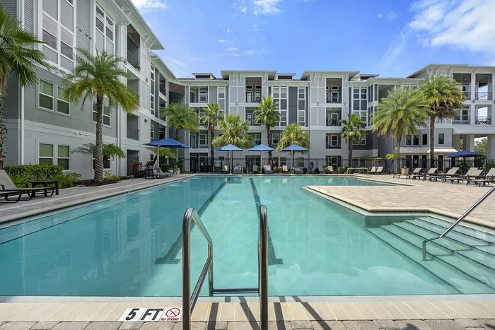 Modern apartment complex with a luxurious pool surrounded by palm trees and spacious lounging areas under blue umbrellas.