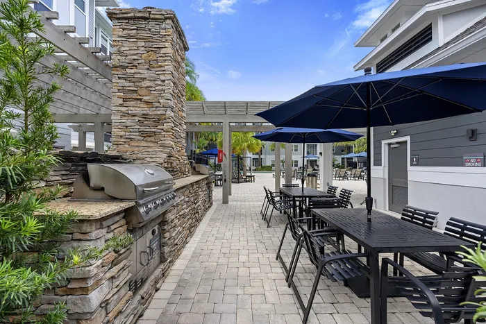 Charming outdoor patio with stone grill, dining set under blue umbrellas, and lush greenery, perfect for entertaining.