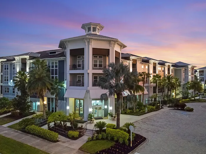 Elegant apartment building at sunset with lush landscaping and modern design, featuring illuminated entryways and palm trees.