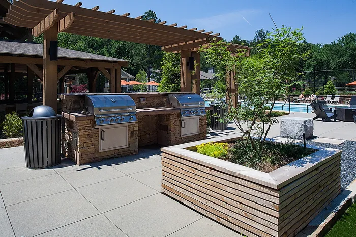 Outdoor luxury grilling area with modern stainless steel barbecues, surrounded by lush greenery and a resort-style swimming pool.
