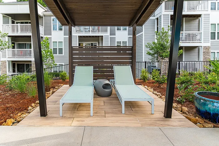 Chic outdoor patio with two sleek lounge chairs under a stylish pergola, surrounded by lush greenery and modern apartment balconies in the background.