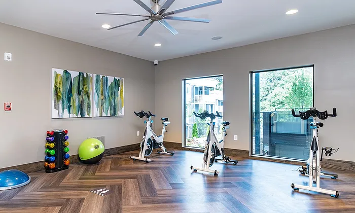 Bright and modern apartment gym with spin bikes, free weights, and stylish wooden flooring, complemented by abstract artwork and large windows offering lush greenery views.