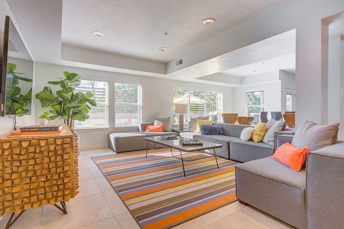 Bright and modern living room featuring a large L-shaped sofa, colorful accent pillows, and a vibrant striped rug, with ample natural light from large windows and stylish decor.