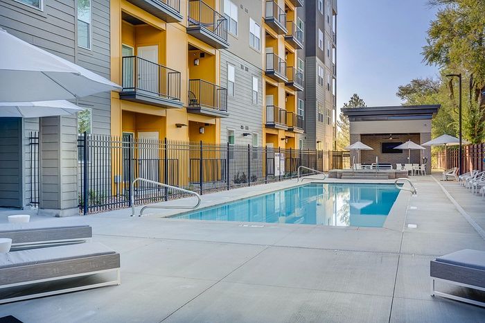 Apartments Near TRAX Salt Lake City, UT - ViA - Resort-Style Swimming Pool With Clear Water, Spacious Sundeck, Lounge Chairs, Shade Umbrellas, and Multi-Story Apartment Building Views.
