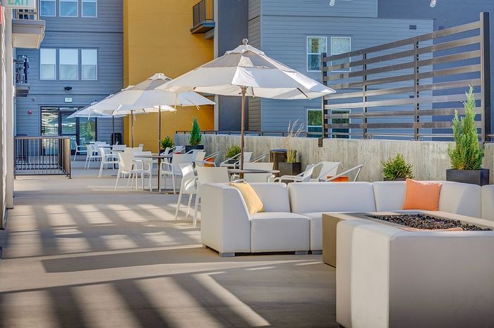Apartments With EV Charging in Salt Lake City, UT - ViA - Outdoor Lounge With Sectional Seating, Fire Pit Table, Patio Umbrellas, Caf&eacute; Tables, and Apartment Building Backdrop.