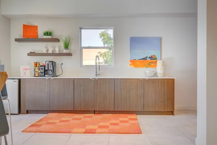 Modern kitchen area with sleek wooden cabinets, vibrant orange rug, and artistic decor. Natural light fills the space, offering a cozy and inviting atmosphere.