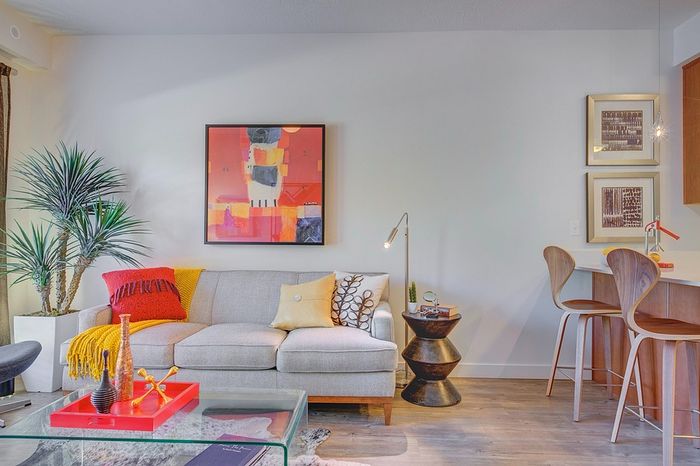 Chic living room with modern decor, featuring a cozy gray sofa adorned with colorful cushions, a striking abstract painting, and a stylish glass coffee table, alongside a sleek breakfast bar with elegant stools.
