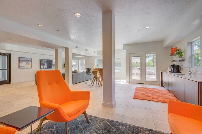 Bright and inviting open-concept living space featuring modern orange accent chairs, a sleek kitchen area with stylish cabinetry, and abundant natural light through large windows.