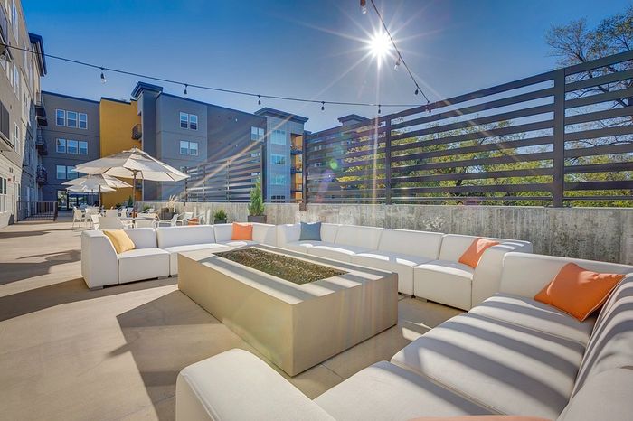 Modern outdoor patio with luxurious white seating, colorful cushions, and a sleek fire pit under a sunny sky.