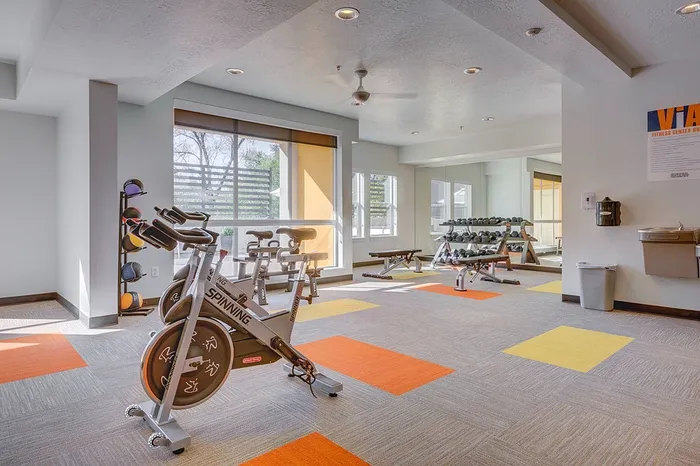 Inviting fitness center with natural light, featuring spinning bikes, colorful exercise mats, and a well-equipped weight rack.