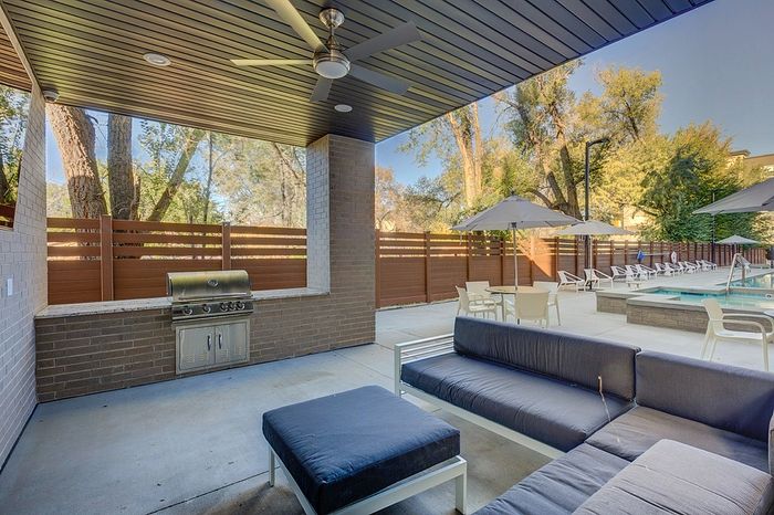 Modern outdoor patio with sleek seating, grill, and cozy poolside area, surrounded by lush trees and umbrellas.