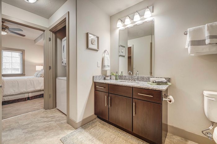 Bright modern bathroom featuring a granite countertop vanity, sleek lighting, and convenient in-unit laundry beside a cozy bedroom.