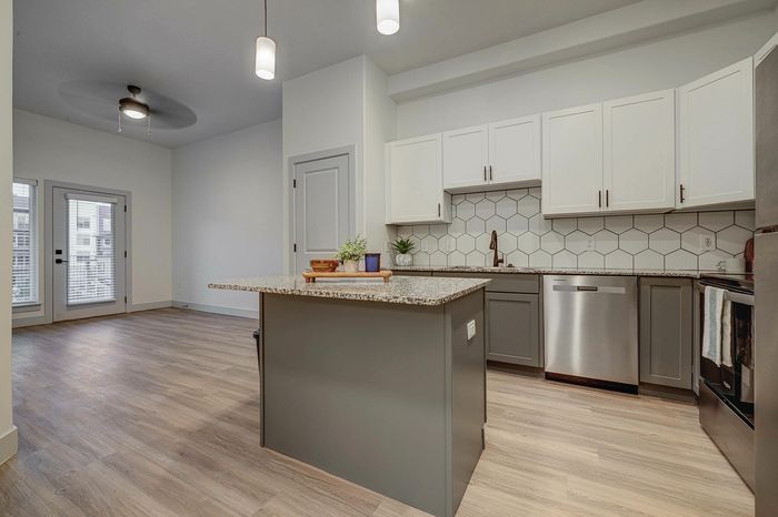 Modern kitchen with sleek granite countertops, stainless steel appliances, and stylish hexagon tile backsplash opening to a bright living area with large windows.