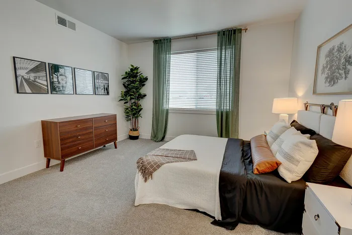Inviting bedroom with modern decor, featuring elegant artwork, a chic dresser, and a cozy bed with stylish pillows, complemented by green curtains and a lush floor plant.