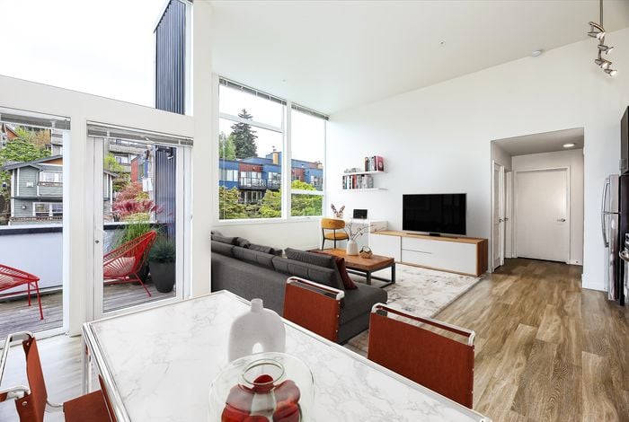 Bright and modern living space with open floor plan, featuring a sleek dining area, cozy gray sofa, large windows opening to a balcony with vibrant red chairs, and stylish wood flooring.
