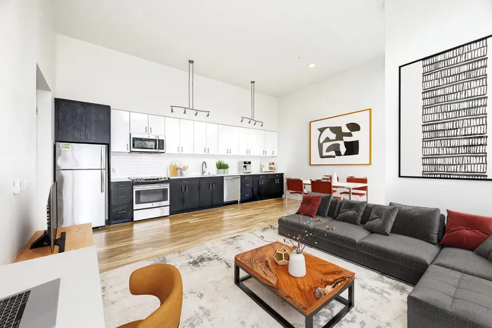 Modern open-concept living area showcasing a sleek kitchen with stainless steel appliances, cozy gray sectional sofa, and elegant dining space adorned with contemporary art.